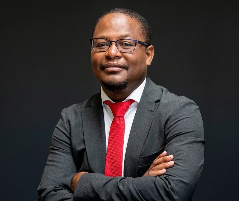 Professional head-and-shoulders portrait of a business executive wearing a dark suit, white shirt and red tie, standing with folded arms against a dark background.